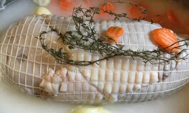 Préparation de la cuisson tête de veau cocotte. Le veau est ficelé, entouré de carottes et de thym dans un bouillon.