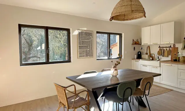 Comedor y cocina luminosos con mesa de madera, sillas de mimbre y tapizadas, y ventanas de cristal térmico que dejan ver el exterior.