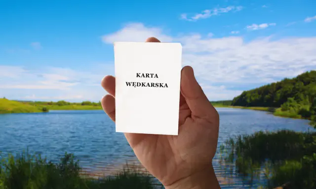 Dłoń trzyma białą kartkę z napisem "KARTA WĘDKARSKA" na tle jeziora i lasu.