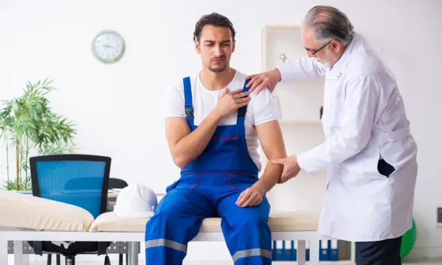 Un ouvrier en bleu de travail consulte un médecin pour une possible maladie professionnelle de l'épaule. Le médecin examine son bras.