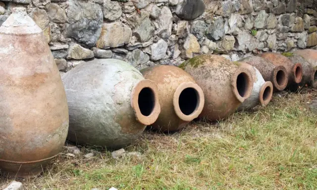 Antiguas ánforas de vino yacen en la hierba, algunas cubiertas de musgo, contra un muro de piedra.