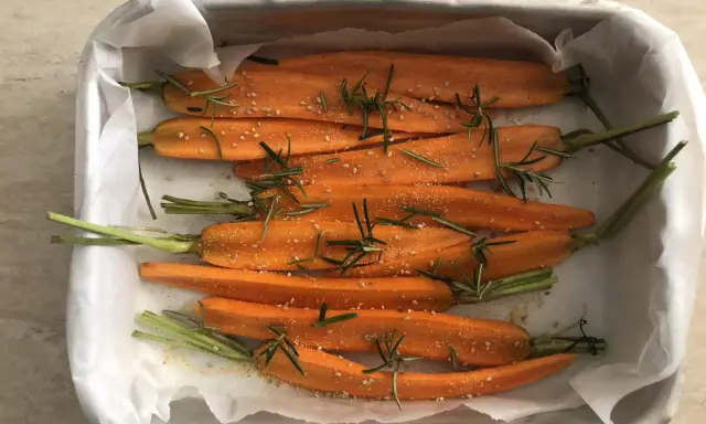 Carote al forno, tagliate a metà, condite con rosmarino e semi di sesamo, pronte per essere infornate.