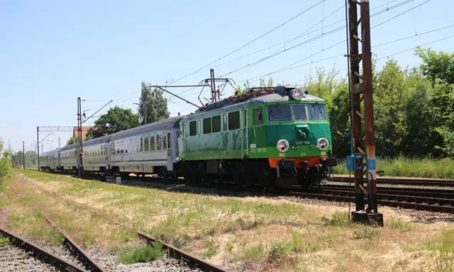 Zielony pociąg EU07-052 z wagonami mija tory w słoneczny dzień. To widok z wieluń trasa.