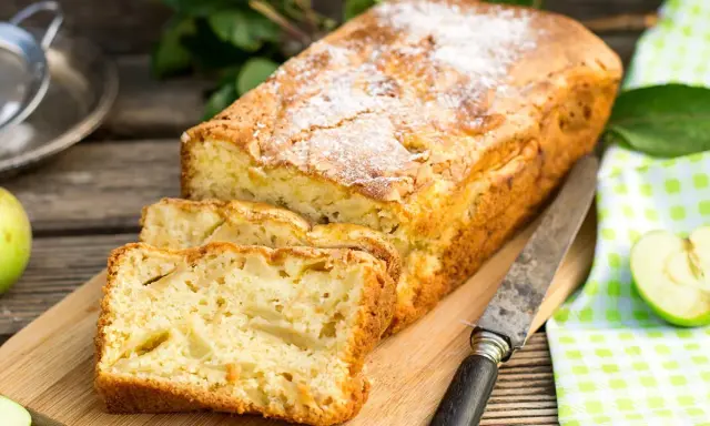 Szybkie ciasto z jabłkami, posypane cukrem pudrem, pokrojone na kromki, leży na desce obok jabłek i noża.