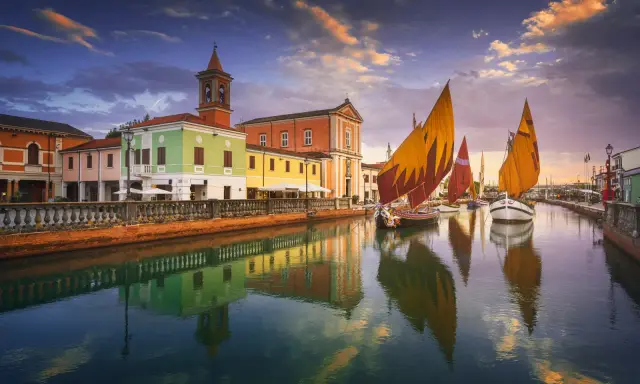 Navi a vela colorate navigano sul canale di Cesenatico al tramonto, con edifici vivaci e riflessi sull'acqua. Meglio Cervia o Cesenatico? Cesenatico offre questo spettacolo.