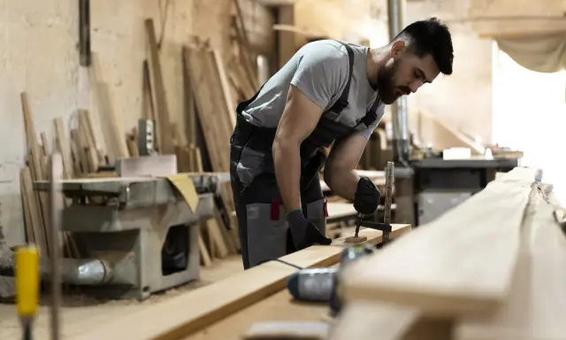 Carpintero ajustando una pieza de madera con una prensa. En el taller, se aprecian varios tablones y maquinaria, listos para crear acabados de madera perfectos.
