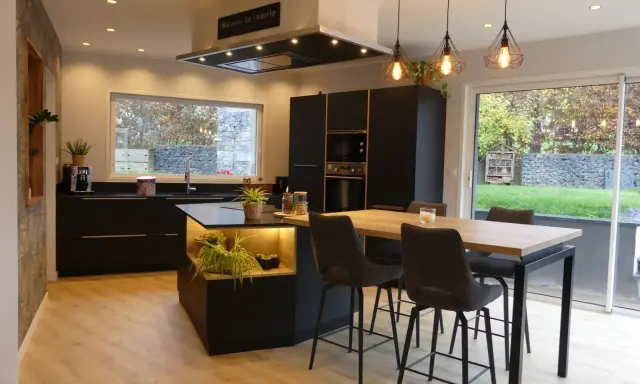 Cuisine noir et bois, mur gris texturé. Îlot central avec éclairage, table haute et tabourets. Fenêtre donnant sur jardin.