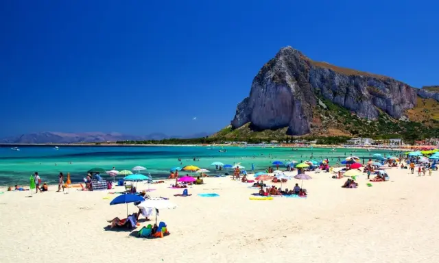 Spiaggia affollata con ombrelloni colorati, gente che si gode le vacanze al mare con bambini, acque turchesi e una montagna imponente sullo sfondo.