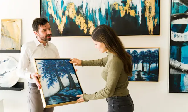 A man and woman admire a painting of blue trees at an art exhibition.
