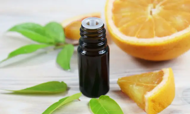 Flacon d'huile essentielle de citrus sinensis, entouré de feuilles vertes et de morceaux d'orange fraîche sur fond de bois clair.