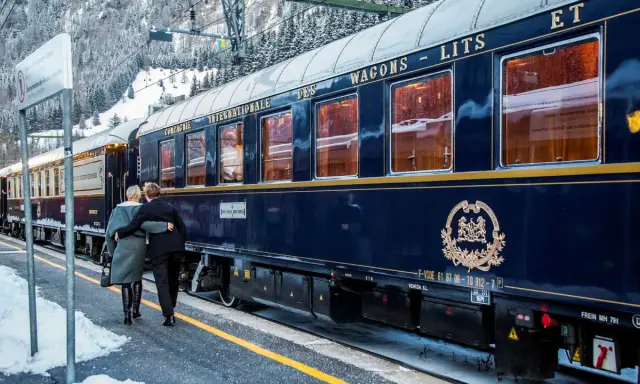 Ett par omfamnar varandra vid en snötäckt station, redo för en resa med Orientexpressen. Tågets mörkblå vagnar med gyllene detaljer vittnar om dess klassiska orientexpressen rutt.