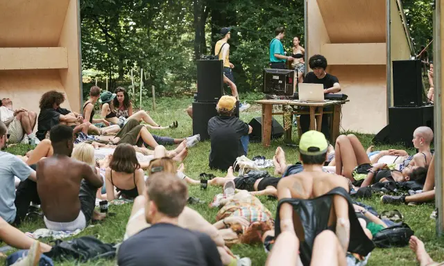 Un gruppo di persone rilassate su un prato in un **boutique festival**, con un DJ che suona musica in sottofondo.