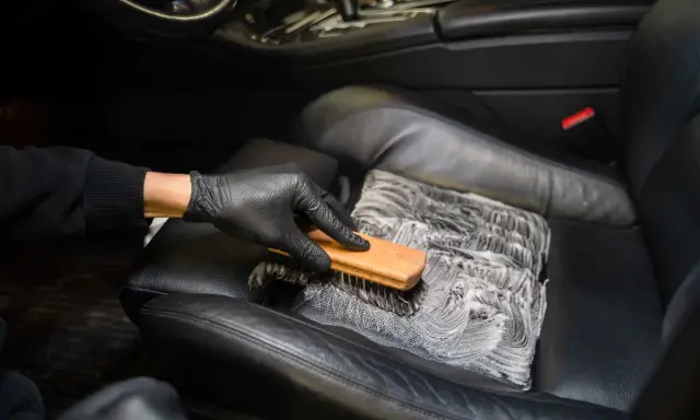 Mano enguantada cepilla asiento de cuero negro con espuma, mostrando cómo limpiar asientos de cuero para un coche impecable.
