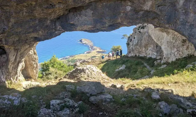 Vista desde el "ojo del diablo", una cueva natural, hacia un paisaje costero con un istmo y el mar azul. Un grupo de personas observa el panorama.