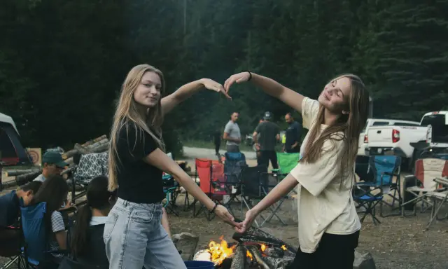 Dwie dziewczyny tworzą serce z dłoni na tle ogniska i lasu. To idealny okres na wakacjach, pełen radości i wspólnych chwil.