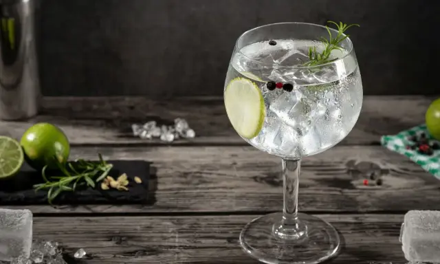 Orzeźwiający gin and tonic z plasterkiem limonki, gałązką rozmarynu i ziarnami pieprzu, na drewnianym blacie.