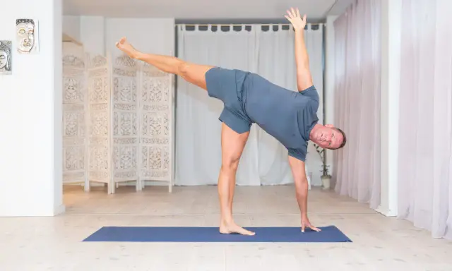 Man utför en dynamisk yoga-position på en blå matta i ett ljust rum med vita draperier och en vikbar rumsavdelare.