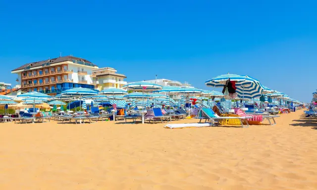 Słoneczny dzień na plaży w Lido di Jesolo. Rzędy leżaków i parasoli w niebiesko-białe pasy czekają na plażowiczów.