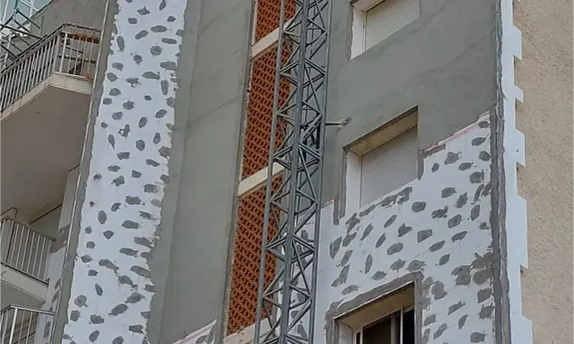 Proceso de como aislar una pared ya construida con placas de poliestireno y mortero. Se observa una grúa de obra.