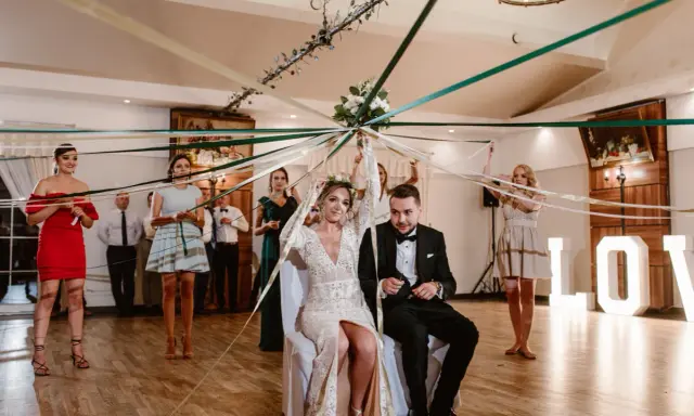 Panna młoda i pan młody siedzą pod baldachimem z kolorowych wstążek, co zamiast oczepin. Goście trzymają wstążki, tworząc magiczną atmosferę.
