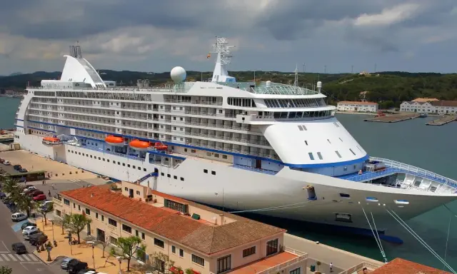 Grande nave da crociera attraccata nel porto Mahon, con edifici storici e colline verdi sullo sfondo.
