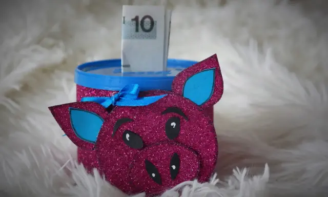 Urocza skarbonka diy w kształcie różowej świnki z brokatem, z niebieskimi uszami i kokardką. W środku widać banknot 10 zł.