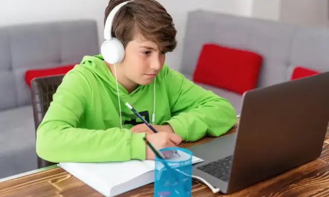 Chłopiec w słuchawkach uczy się zdalnie, notując w zeszycie. Zastanawia się, ile kosztują studia, gdy widzi ceny kursów online.