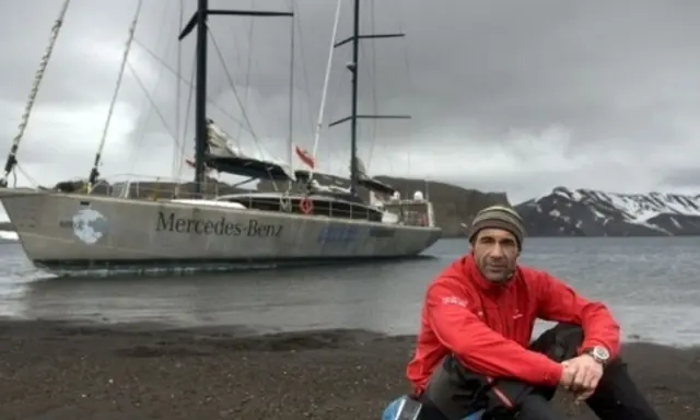 Mike Horn, l'explorateur, pose près de son bateau "Mercedes-Benz" sur une plage rocheuse, avec des montagnes enneigées en arrière-plan.