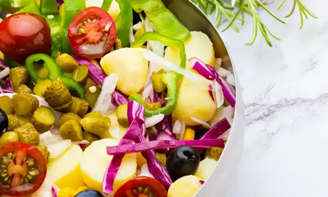 Ensalada campera vibrante con patatas, tomates, pepinillos, pimientos verdes, cebolla morada y aceitunas negras en un bol metálico.