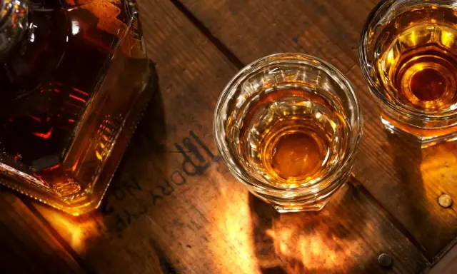 Butelka i dwa kieliszki z bursztynowym płynem, przypominającym brandy lub whisky, stoją na drewnianym blacie.