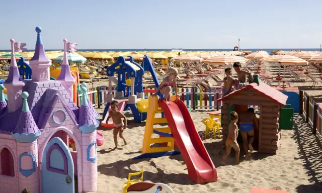 Bambini giocano in un'area giochi colorata sulla sabbia, con castello e scivoli, mentre ombrelloni gialli e lettini creano un'atmosfera da **sunday beach Rimini**.