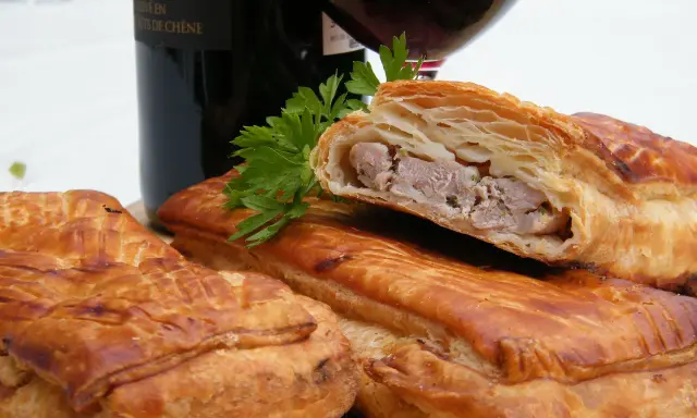Pâté en croûte, une spécialité lorraine savoureuse, avec une garniture de viande visible. Une bouteille de vin rouge et du persil complètent la scène.