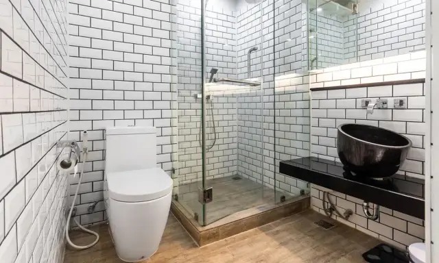 Baño moderno con azulejos blancos y negros. El suelo de madera se ve primero, luego los azulejos. Inodoro, ducha de cristal y lavabo negro.