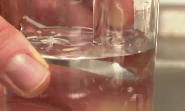 Main d'une personne tenant un verre d'eau potable à Poitiers. L'eau est claire et reflète la lumière.