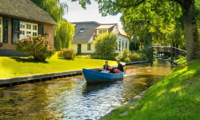 Ein Paar fährt mit einem blauen Boot durch einen Kanal im "Klein Venedig Holland". Reetgedeckte Häuser und grüne Ufer säumen den Weg.