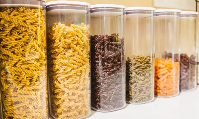 Contenitori trasparenti pieni di pasta a forma di farfalline, di vari colori: gialle, marroni, verdi e arancioni.