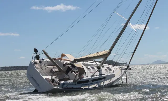 Un voilier échoué, incliné sur le côté, témoigne d'une possible collision bateau. Les vagues s'écrasent sur sa coque blanche sous un ciel bleu.