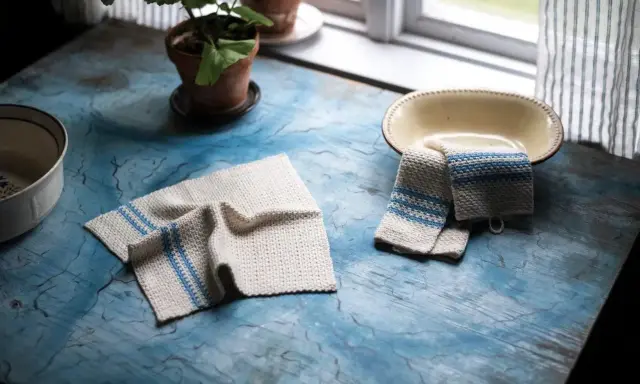 Två virka handdukar med blå ränder ligger på ett blått bord. En krukväxt och en skål syns också.