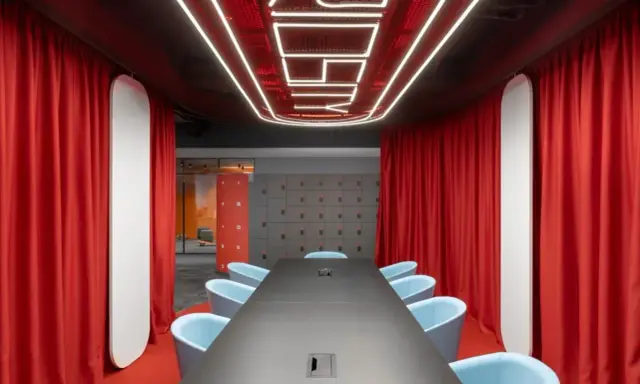 Aménagement salle de réunion moderne avec table noire, fauteuils bleus, rideaux rouges et éclairage néon au plafond.