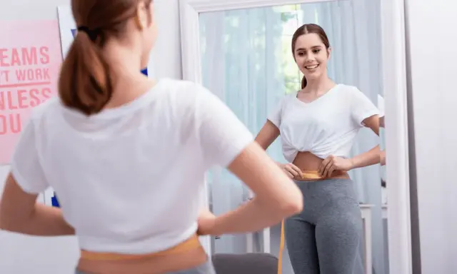 Mujer se mide la cintura con cinta métrica, sonriendo al verse en el espejo. No hay evidencia de que el diu de cobre engorde.