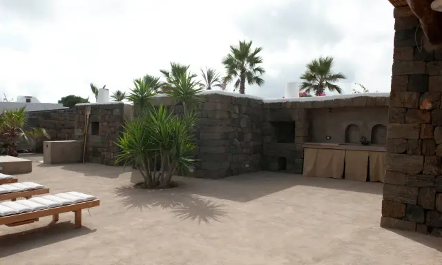 Un cortile in cemento trasformato con piante rigogliose, un barbecue in pietra e comodi lettini. Idee per come abbellire un cortile in cemento.
