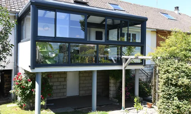 Une extension véranda toit plat moderne, avec de larges baies vitrées, s'intègre parfaitement à la maison. Un fauteuil vert est visible à l'intérieur.