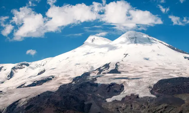 Elbrus, najwyższy szczyt Rosji, wznosi się dumnie pod błękitnym niebem z kłębiastymi chmurami. Lodowce spływają po jego zboczach.