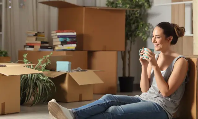 Une femme boit une tasse de café, entourée de nombreux cartons de déménagement.