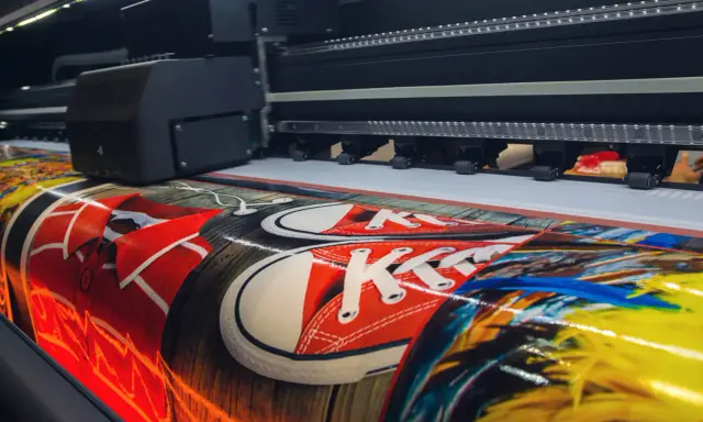 Wielki plakat reklamowy z czerwonymi trampkami i kolorowymi piórami powstaje na drukarce wielkoformatowej.