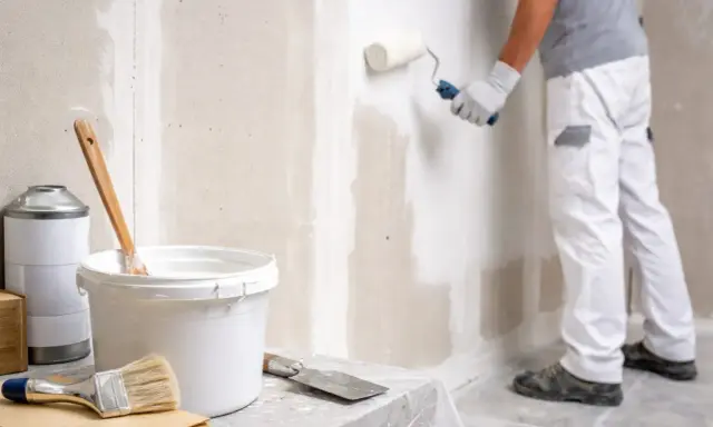Un peintre applique une couche de peinture sur un mur en Fermacell. Matériel de peinture à proximité.
