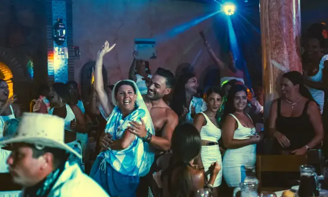 Gente celebrando y bailando en una fiesta, con luces de discoteca y un ambiente festivo. Parece una despedida de soltero con juegos y mucha alegría.