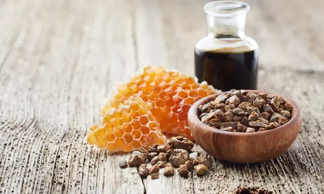 Propolis i pyłek pszczeli w drewnianej miseczce, obok plastra miodu i buteleczki z ciemnym płynem, na drewnianym tle.