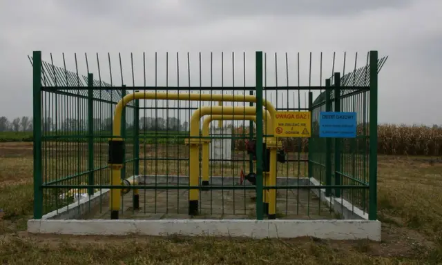 Ogrodzony obiekt gazowy z żółtymi rurami. Tablice ostrzegawcze informują o zagrożeniu. Widać pola uprawne w tle, co sugeruje odległość budynku od gazociągu średniego ciśnienia.