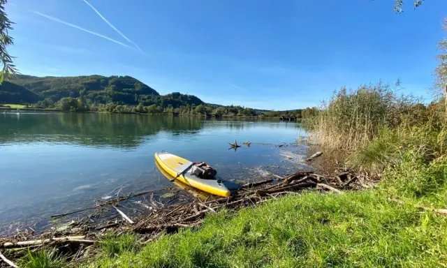 Gelbes SUP-Board liegt am Ufer bereit für SUP Touren Bayern. Ruhiger See, bewaldete Hügel und blauer Himmel.